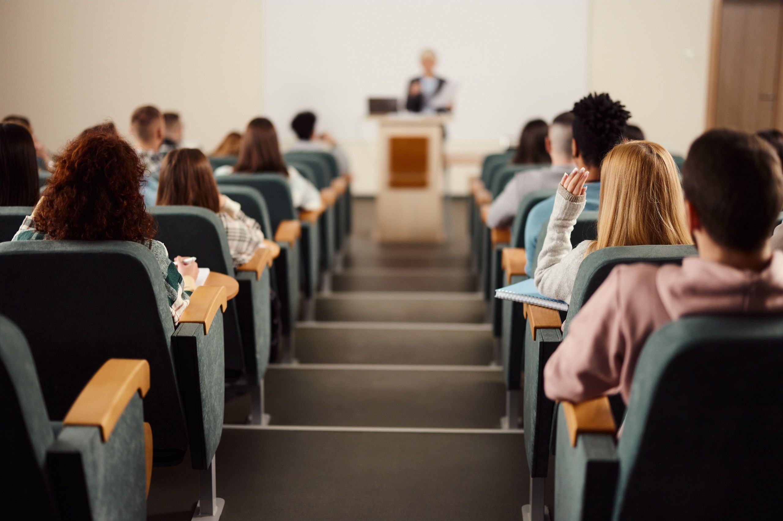 studenci siedzący w rzędach amfiteatralnej sali wykładowej, jedna ze studentek podnosi rękę podczas wykładu, w tle wykładowca przy mównicy