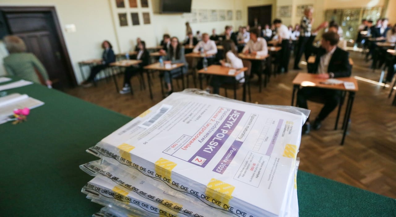 Stos zapakowanych arkuszy egzaminacyjnych z języka polskiego na maturze, w tle zdający piszący egzamin w sali