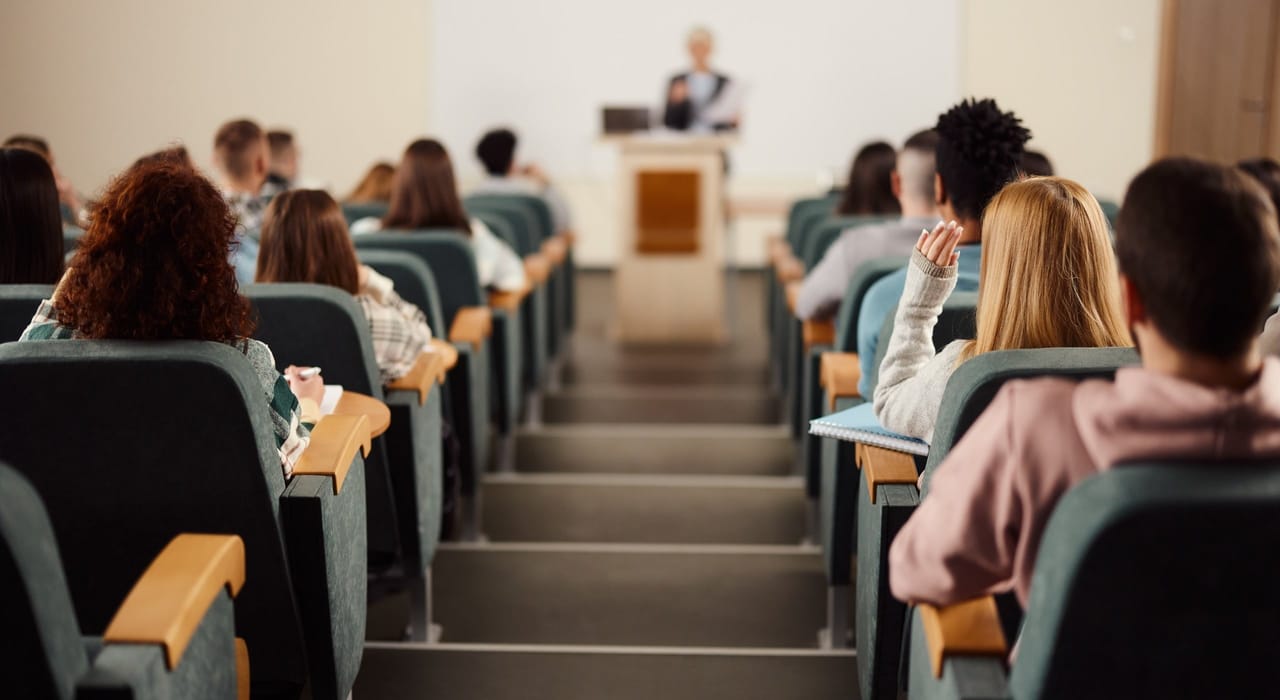 studenci siedzący w rzędach amfiteatralnej sali wykładowej, jedna ze studentek podnosi rękę podczas wykładu, w tle wykładowca przy mównicy