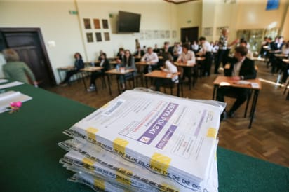 Stos zapakowanych arkuszy egzaminacyjnych z języka polskiego na maturze, w tle zdający piszący egzamin w sali