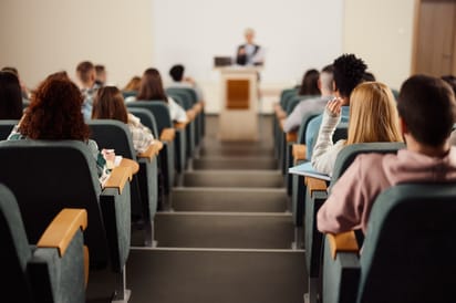 studenci siedzący w rzędach amfiteatralnej sali wykładowej, jedna ze studentek podnosi rękę podczas wykładu, w tle wykładowca przy mównicy