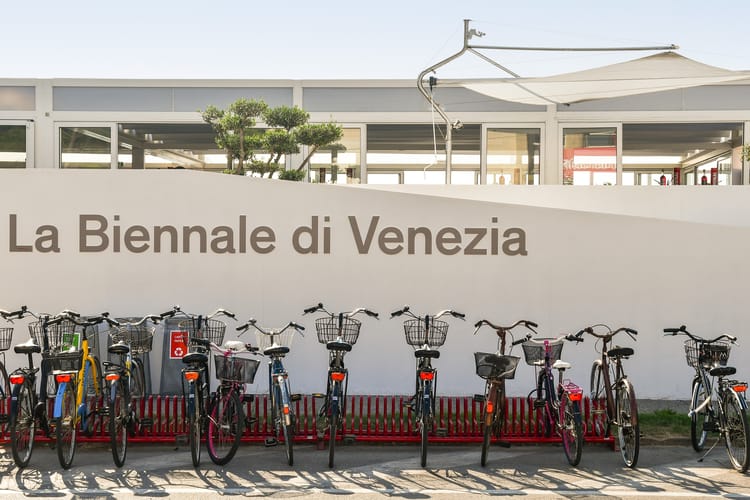Rząd rowerów zaparkowanych przed ścianą z napisem „La Biennale di Venezia”.