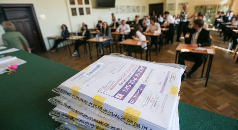 Stos zapakowanych arkuszy egzaminacyjnych z języka polskiego na maturze, w tle zdający piszący egzamin w sali